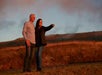 A couple stands outdoors at sunset. The woman points towards the horizon, and the man stands beside her. The ground and sky are bathed in warm, orange light.