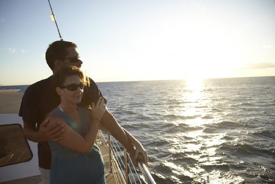 Two people wearing sunglasses stand on a boat deck, looking at the sunset over the ocean.