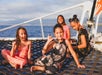 Four girls sit and pose on the net of a boat, with the ocean and sky in the background on a sunny day.