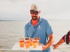 A man in a blue shirt and cap holds a tray with six iced drinks on a boat, with water and a cloudy sky in the background.