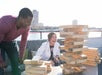 Two people play giant Jenga on a rooftop with city buildings in the background. The woman is smiling, while the man watches the tower closely.