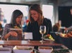 Two women selecting food at a buffet. One is holding a plate and pointing at dishes, while the other smiles, holding a plate. A glass of white wine is on the table.