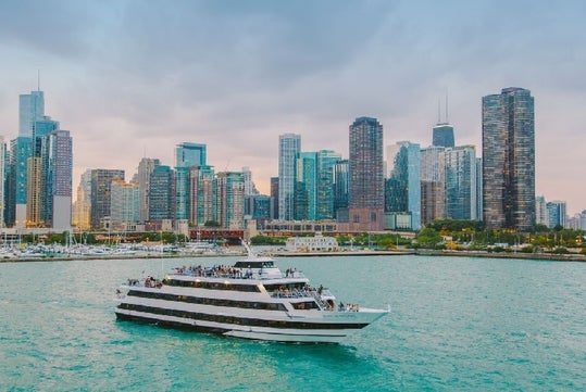 A white multi-deck boat sails on green water against a backdrop of a modern city skyline with tall buildings under a cloudy sky.