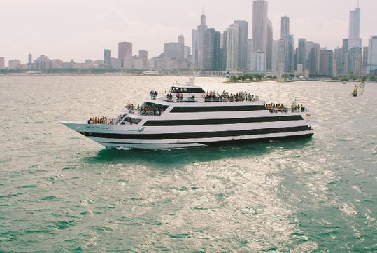 A large white and black-striped tour boat on a city waterfront, with a skyline in the background and people on board.