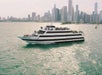 A large white and black-striped tour boat on a city waterfront, with a skyline in the background and people on board.