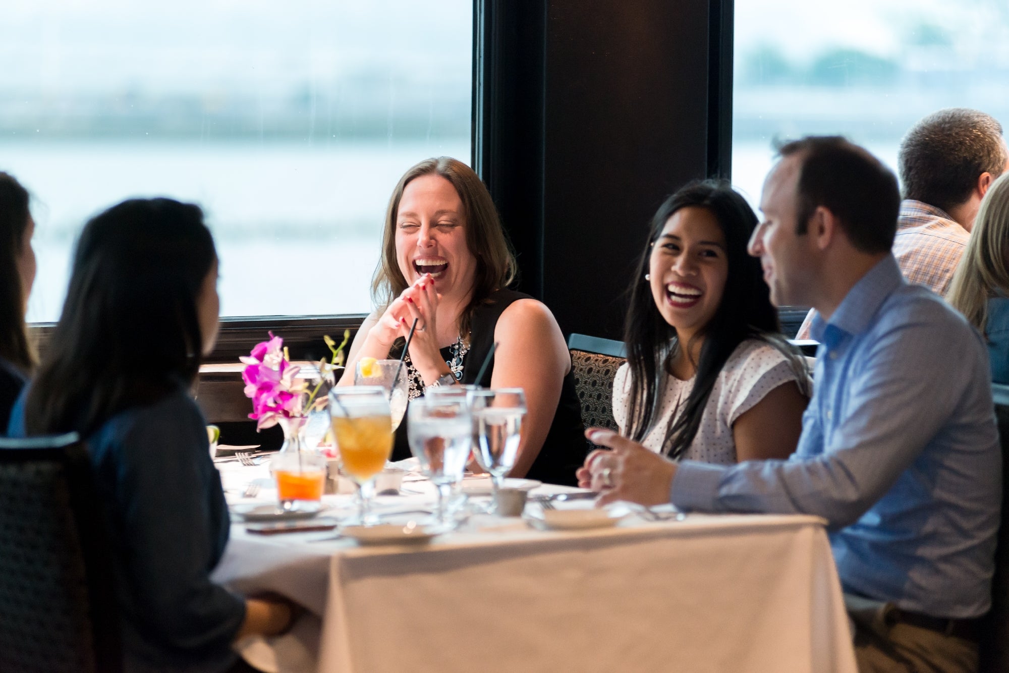 Four people sit at a restaurant table by a window, engaged in conversation and laughing. There are drinks, flowers, and tableware on the table.