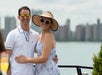 A man in a white polo shirt and a woman in a sunhat and dress stand together outdoors by the water, with a city skyline in the background.