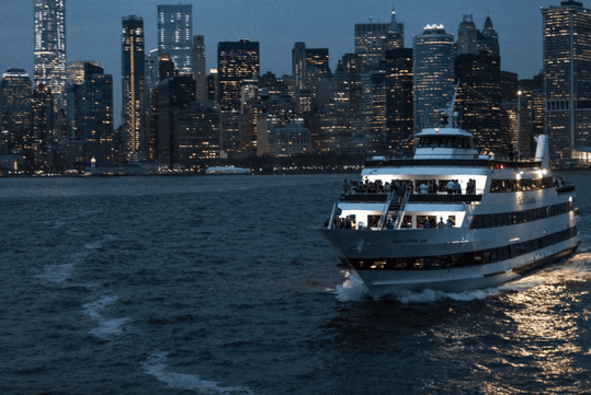 A cruise boat with people standing on it's deck at night with the city of New York in the background.