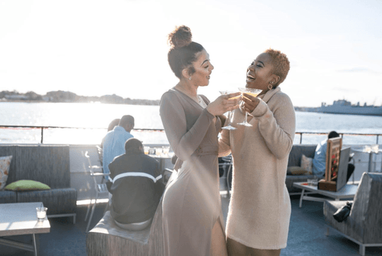 Two women in beige dressed standing together and laughing while cheering their drinks together on the Spirit of Norfolk Signature Brunch Cruise.
