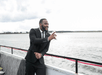 A man in a suit holding a glass smiling and standing near the railing of a cruise boat with water in the background.