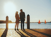A man and woman holding hands and standing on a pier with the sun setting in front of them and several sailboats in the water.