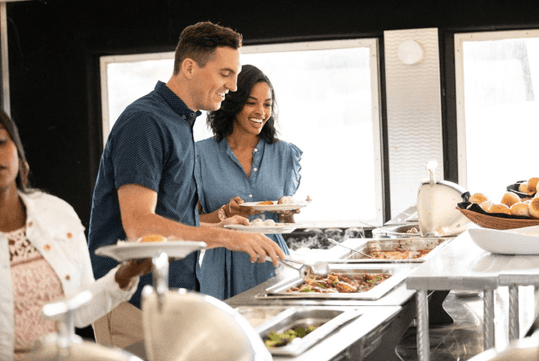 A man and woman standing in front of a tiered buffet smiling with plates in their hands and large windows behind them letting light it.