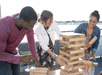 Three friends smiling and laughing while playing giant Jenga on the deck of a cruise boat with the sun shining on them and water in the background.