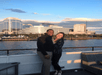 A man and woman standing close together posing for a photo on the deck of a boat with the city of Norfolk behind them at sunset.