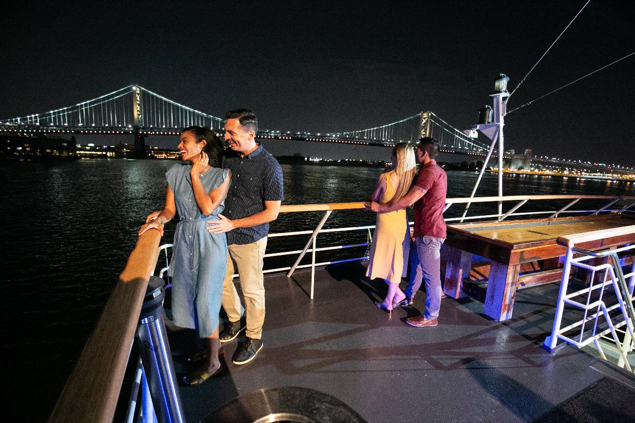 Two couples stand on a boat deck at night with a lit bridge in the background. One couple embraces while the other gazes outward.