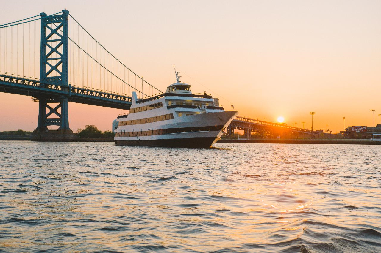 A large boat sails on a river during sunset, with a suspension bridge and cityscape visible in the background.