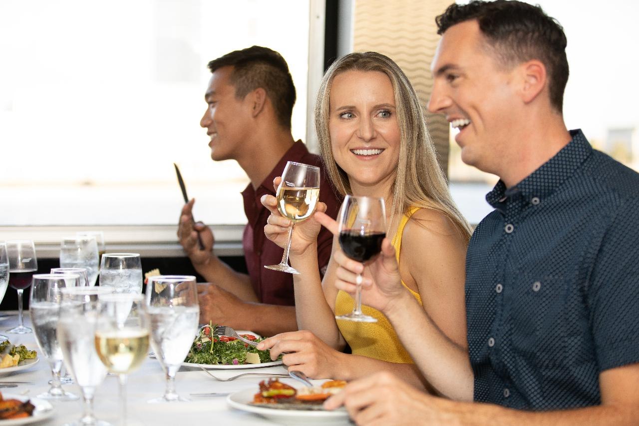 Three people sit at a table in a restaurant, holding wine glasses and eating salads. They are engaged in conversation and smiling.