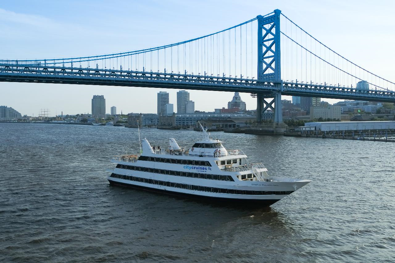 A city cruise boat sails under a large suspension bridge, with an urban skyline in the background and calm water below.