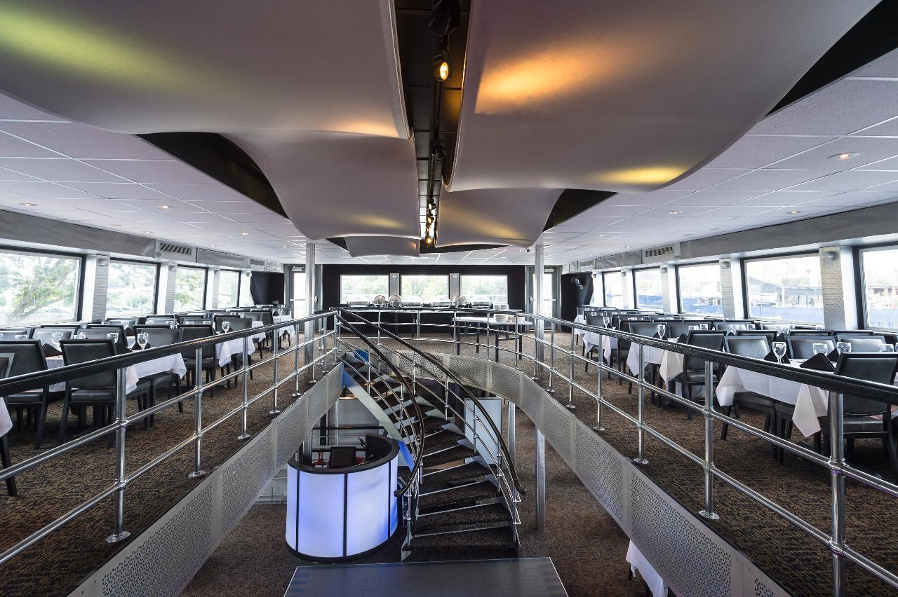 Interior of a modern restaurant with an upper mezzanine floor, featuring tables with white tablecloths, black chairs, and a central staircase with metallic railings.