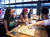 People playing board games at a table in a dimly lit room. A woman smiles at a board game, while others engage in games around her. Drinks are on the table.