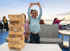 A child holds up a wooden Jenga block triumphantly, standing next to a stacked Jenga tower on a table. Another child stands nearby, with water and people in the background.