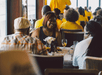 People sitting at a dining table with food and drinks, engaged in conversation. Brightly lit room with others in yellow shirts in the background.
