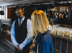 A bartender smiles while conversing with a woman in a bar. Another woman stands behind the bar amidst liquor bottles and glasses.