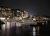 A large boat is illuminated and docked at a marina at night. Brightly lit buildings reflect on the water in the background.