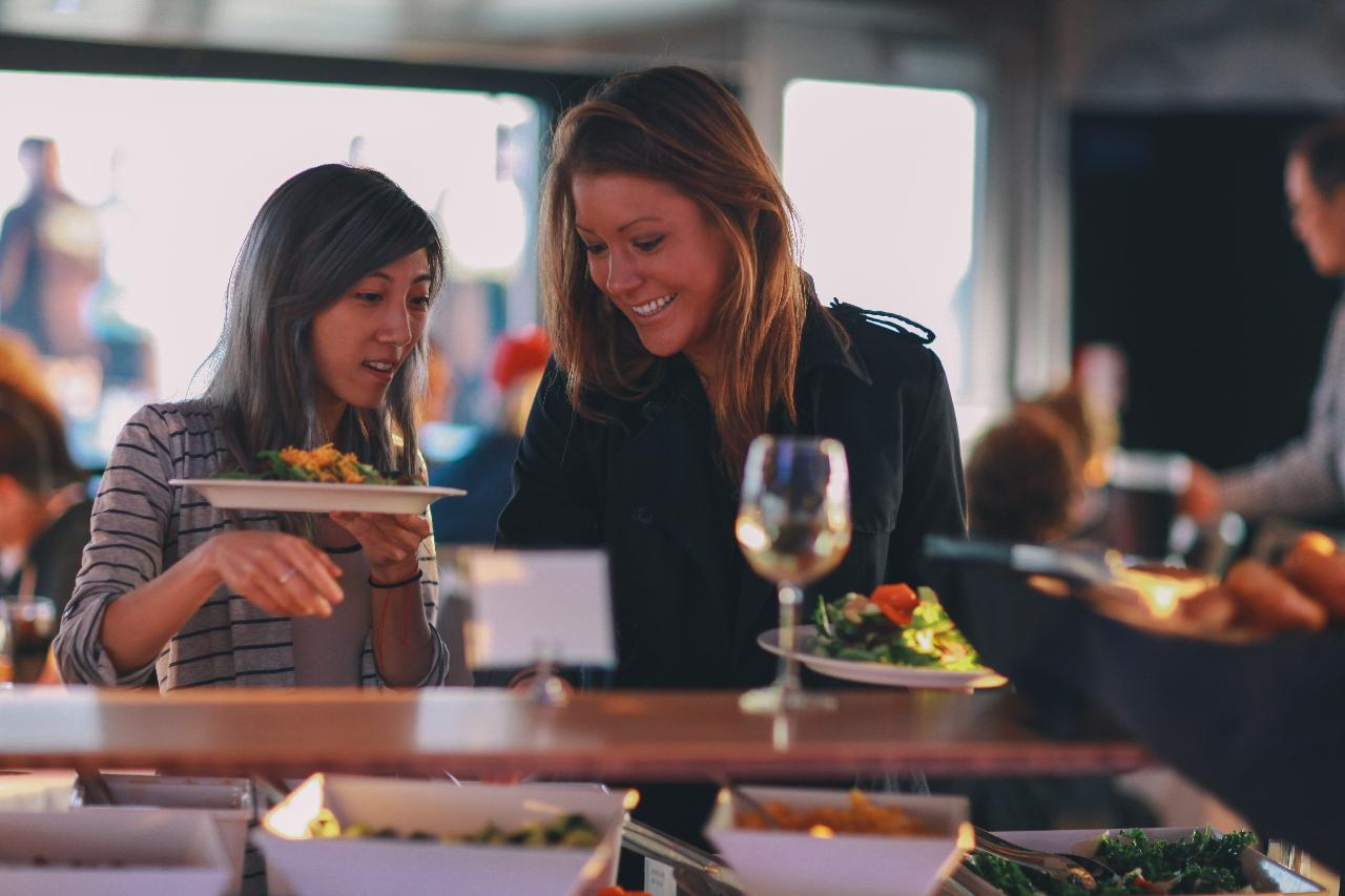 Two women at a buffet, holding plates of food. A glass of white wine is on the counter in front of them.