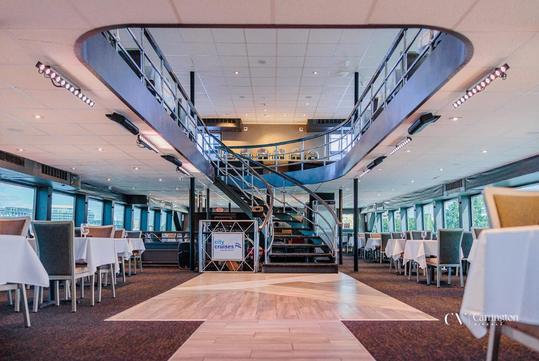 Interior of a cruise ship dining area with a central spiral staircase, white tablecloths, and large windows on either side.