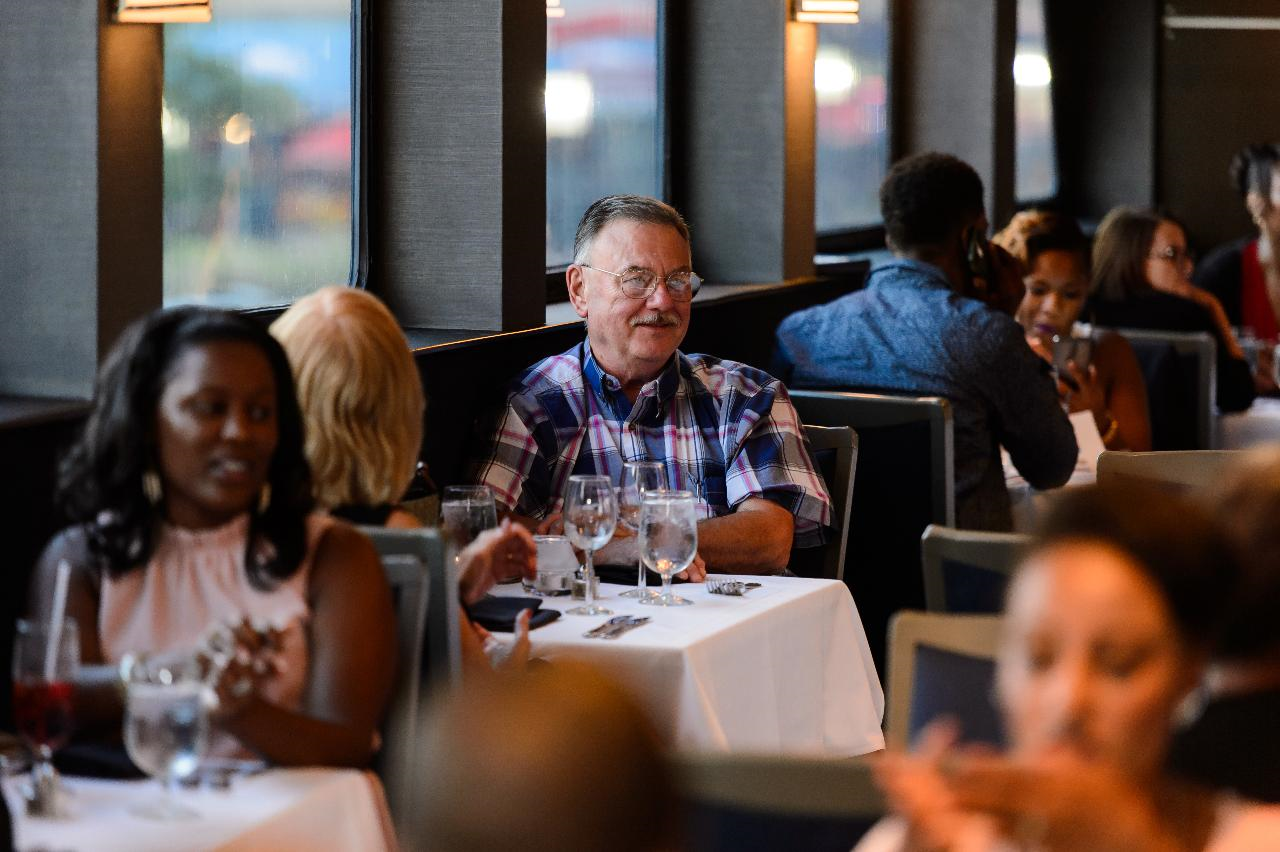 A man in a plaid shirt sits at a restaurant table with drinks, surrounded by other diners in a busy dining area.
