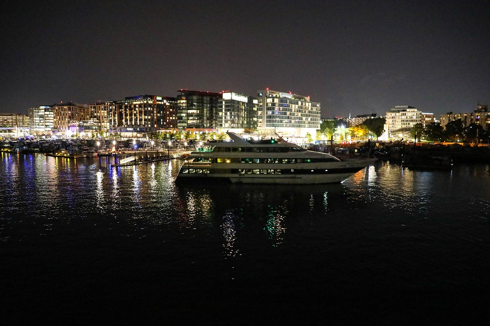 A yacht sails at night on a calm river, with illuminated city buildings in the background.