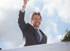 Man in a suit raising a glass and smiling, standing behind a white surface with greenery, against a cloudy blue sky.
