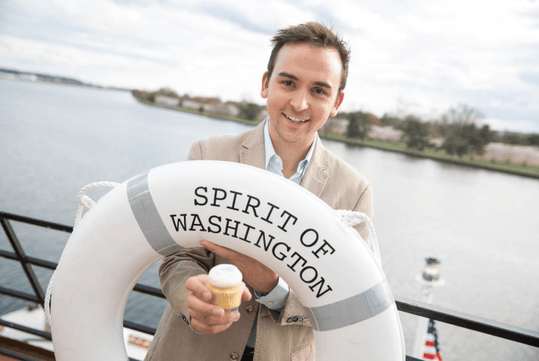Person holding a life ring labeled "Spirit of Washington" and a cupcake, standing on a boat with water and land visible in the background.