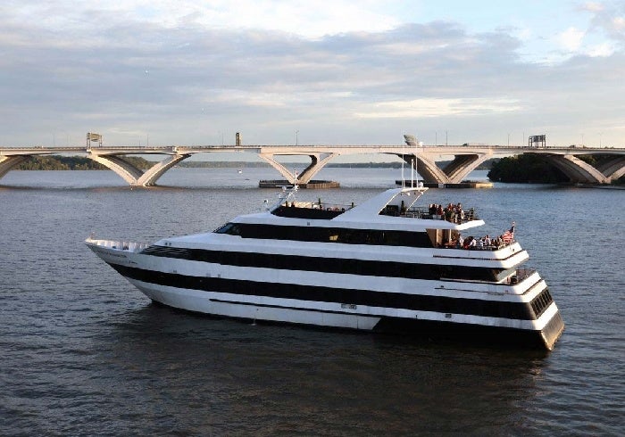 A large white yacht with black stripes sails on a river near a multi-span bridge under a partly cloudy sky.