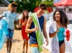 Two teenagers in swimwear, one with a towel draped over his shoulder and the other holding a red tumbler, stand alongside two more teenagers in the background.