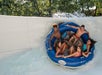Six people ride together on a large blue inflatable raft down a water slide, with splashing water and green trees in the background.