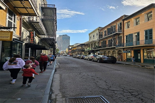 A group of people, including children, walk along a narrow street lined with historic buildings and parked cars under a clear blue sky.