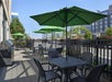 Outdoor patio area with black metal tables and chairs, green umbrellas, a grill, and a black fence, set beside a street and modern buildings.