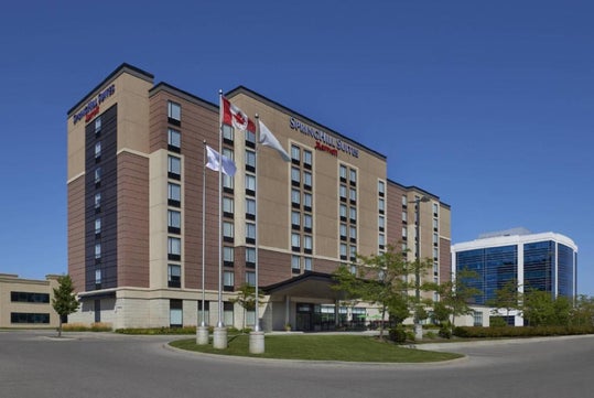 A modern, mid-rise hotel building with a SpringHill Suites sign, three flagpoles, and a glass office building in the background under a clear blue sky.