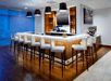 A bar with white chairs and a white counter top with four lights hanging over it and a TV mounted between the shelves on the backwall.