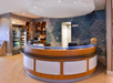 Modern hotel reception area with a circular desk, two monitors, a wall with geometric patterns, and a small shop with snacks and drinks.