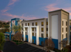 Four-story hotel building with blue accents, large windows, and a main entrance surrounded by palm trees. The sky is clear with a hint of pink during sunset.