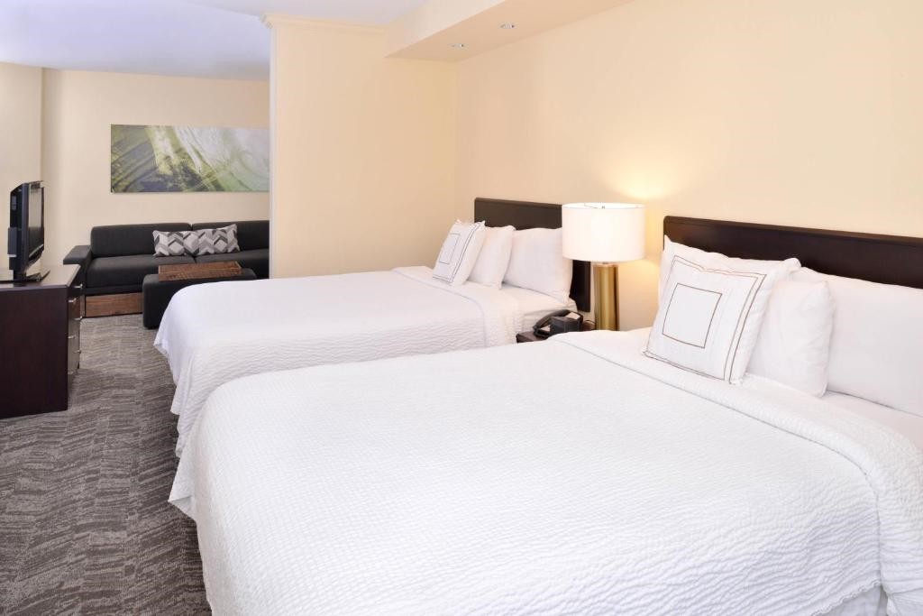 A hotel room with two double beds, white bedding, a sofa, and a TV. The walls are light-colored, and the room has a neutral decor.