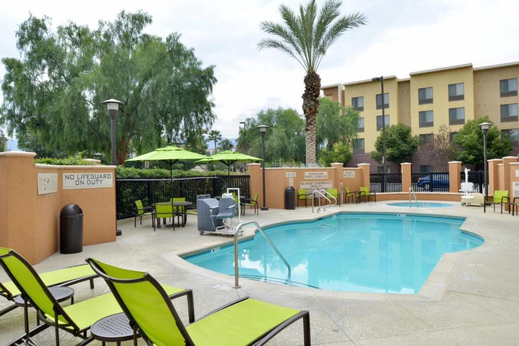 Outdoor hotel pool area with green lounge chairs, umbrellas, and palm trees. Signs indicate no lifeguard on duty. Building and trees in the background.
