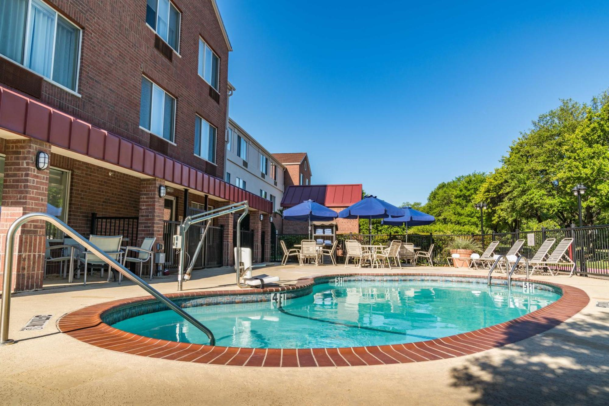 Outdoor hotel pool area with accessible lift, surrounded by lounge chairs and tables with blue umbrellas, next to a brick building under a clear blue sky.