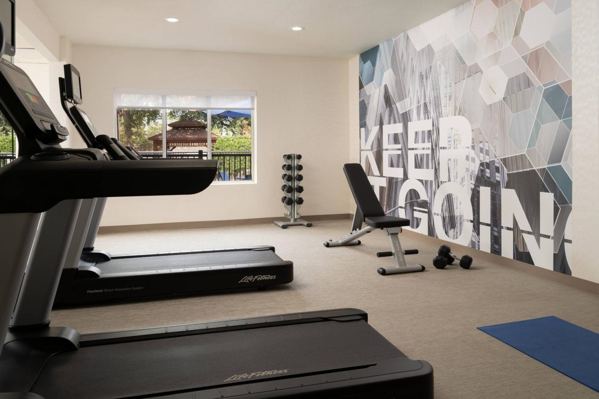 A modern gym room with treadmills, a weight bench, dumbbells, a rack of weights, a blue exercise mat, and a wall mural that says "KEEP GOING.