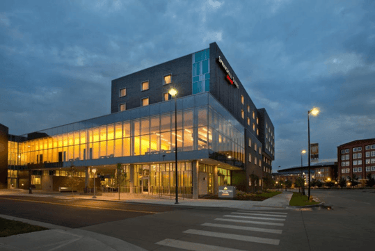 Street view of the SpringHill Suites Denver Downtown at dusk with a cloudy sky over head and warm yellow light visible in the large windows of the hotel.