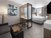 Hotel room with a bed, desk, chair, and sofa. Art on the wall and a TV opposite the bed. Room-divider separates bed and workspace. Large window provides natural light.