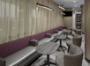 Modern lounge with patterned chairs and tables, purple cushioned benches, textured wall panels, and overhead lighting. A black screen is mounted on the wall in the background.